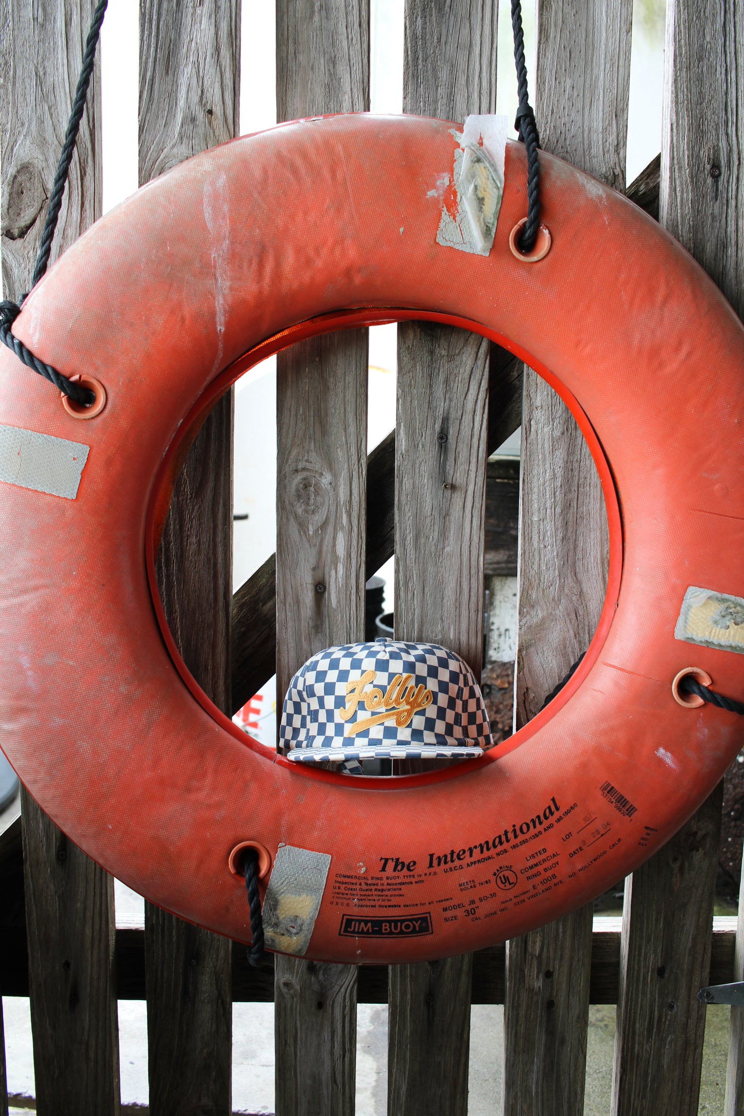 Folly Beach hat. Orange life buoy with a checkered hat inside, hanging on a wooden fence.