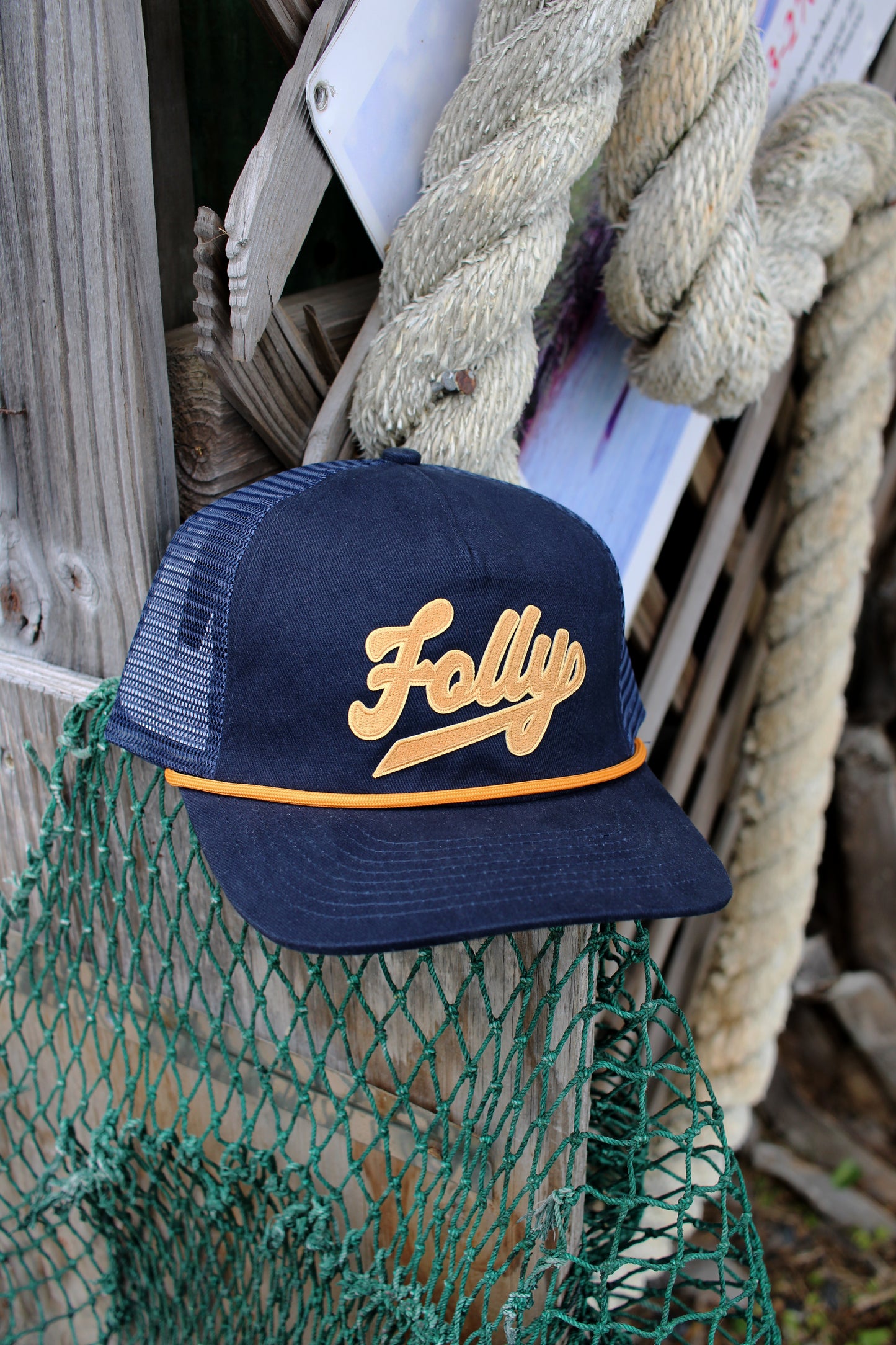 Folly Beach hat. Navy blue cap with 'Folly' logo on a wooden background with ropes and netting.