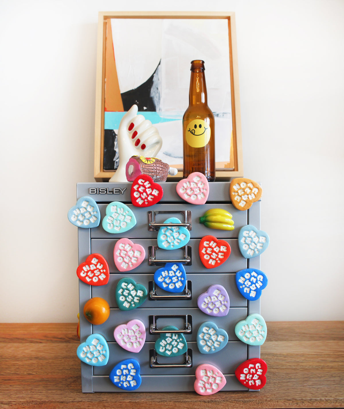 A gray Bisley three-drawer organizer with colorful heart-shaped magnets on the front, filled with pens and stationery, styled on a tabletop with a bottle and fruit props.
