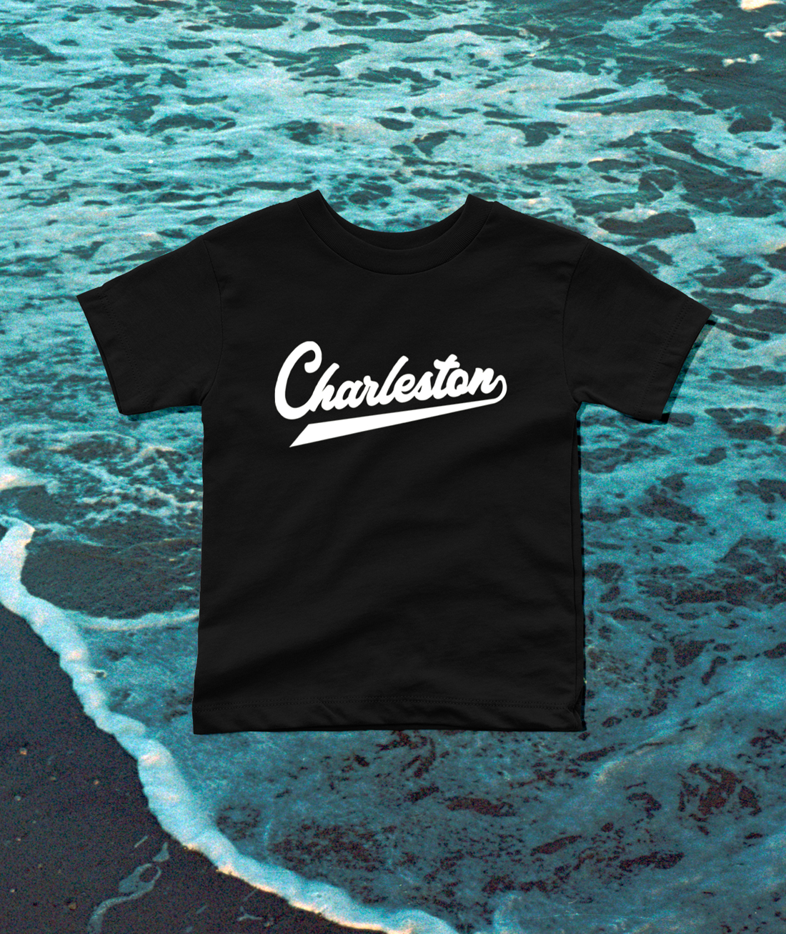 A black short-sleeve T-shirt with the word 'Charleston' printed in white script across the chest, laid flat over an ocean wave background.