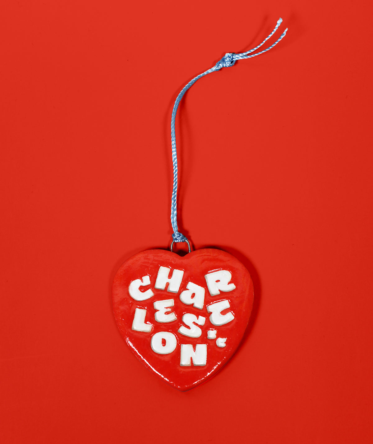 A red heart-shaped hanging ornament with a blue string and white lettering that reads 'CHARLESTON' against a solid red background.