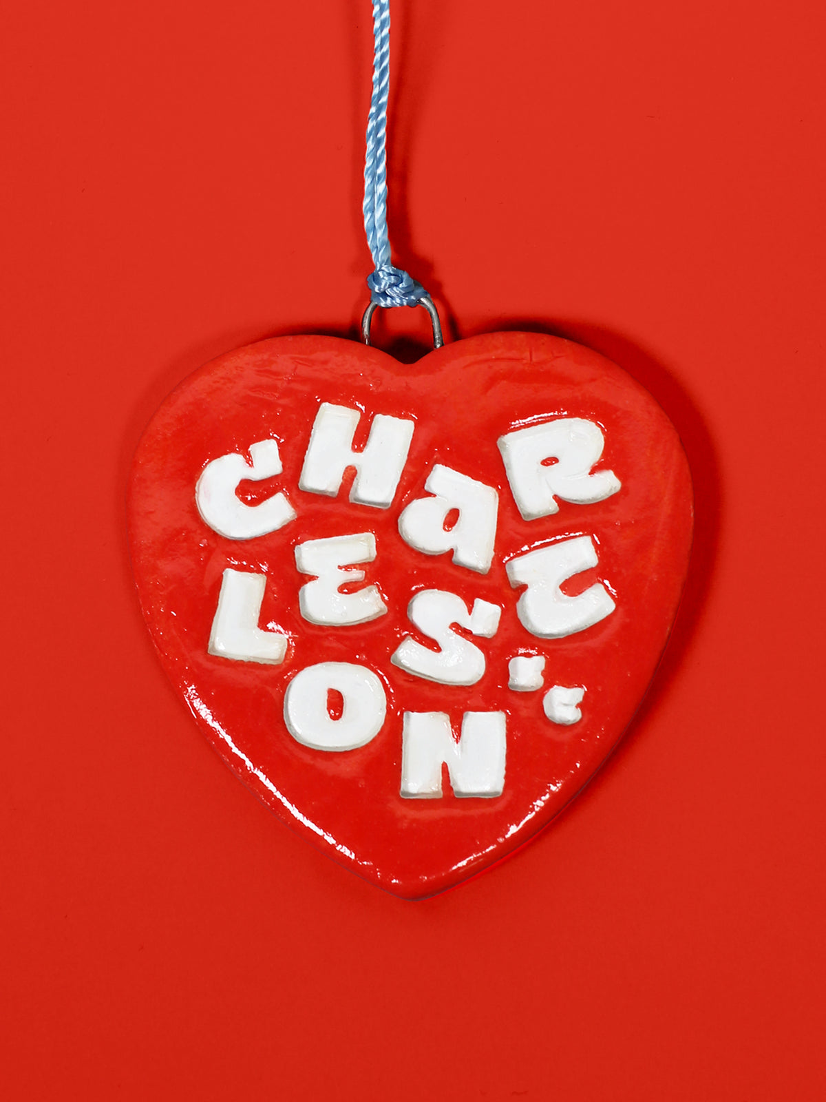 A glossy red heart-shaped hanging ornament with raised white letters spelling 'Charleston' and a blue hanging string, shown against a solid red background.