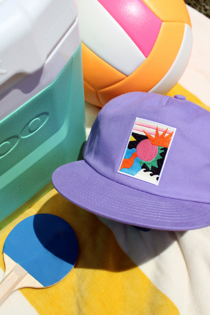 Purple cap with a colorful design, beach ball, and paddle on a sandy surface.