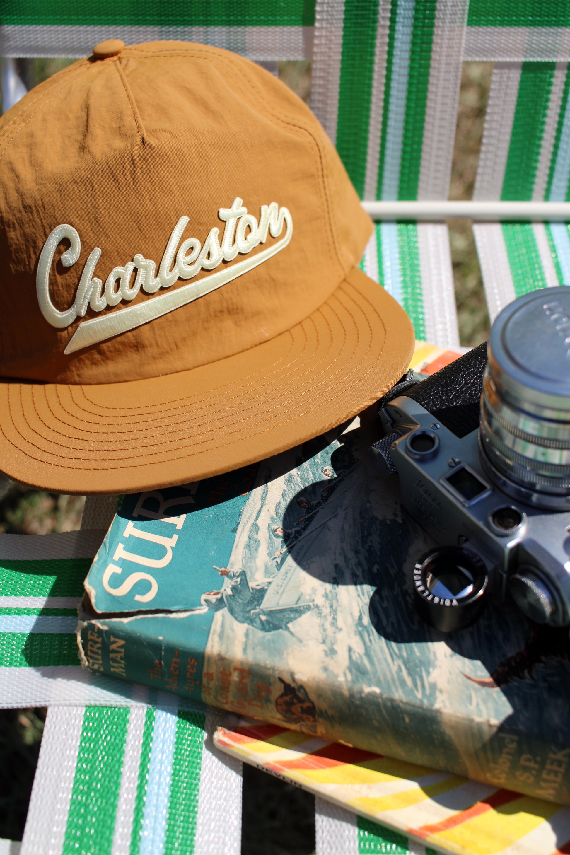 Gold cap with 'Charleston' text on a green and white striped chair with a camera and book.