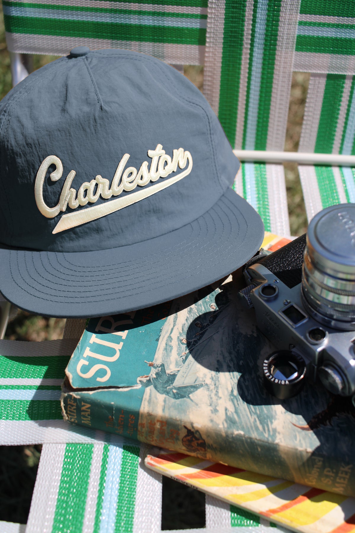 A dark slate blue baseball cap with 'Charleston' embroidered in cream script, styled on a green and white striped patio chair next to a vintage book and camera.
