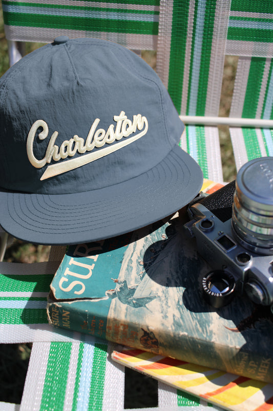 A dark slate blue baseball cap with 'Charleston' embroidered in cream script, styled on a green and white striped patio chair next to a vintage book and camera.