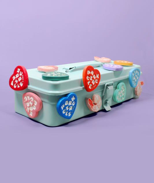 A mint green metal lunchbox decorated with colorful heart-shaped and round buttons on top and sides, set against a solid lavender background.