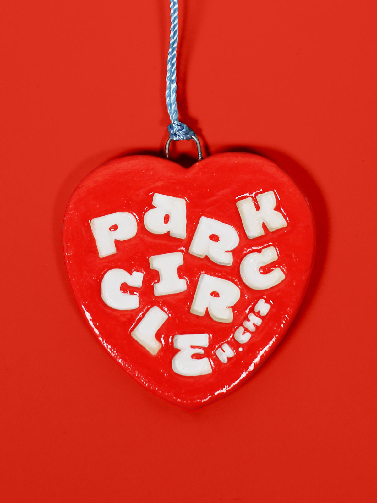 A glossy red heart-shaped hanging ornament with raised white letters spelling 'Park Circle' and a blue hanging string, shown against a solid red background.