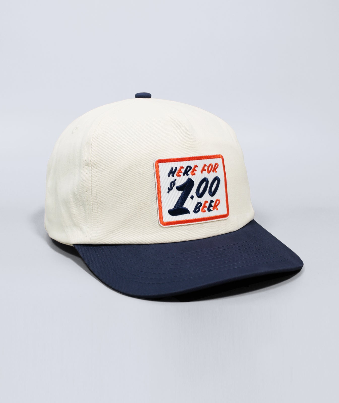 A structured baseball cap with an off-white crown and a dark navy blue flat brim. The front features a rectangular embroidered patch with a red border and the text 'HERE FOR $1.