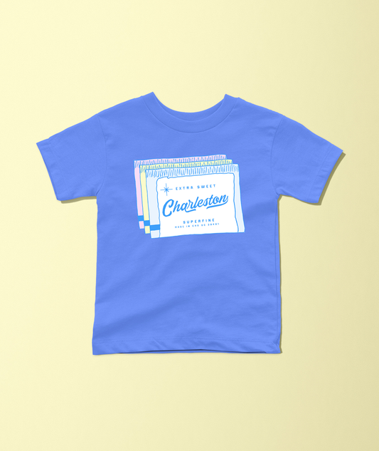 A royal blue short-sleeve t-shirt with a graphic print of a vintage 'Charleston' soda label on the chest, displayed flat against a pale yellow background.