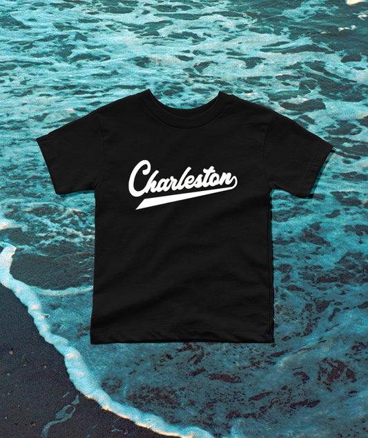 A black short-sleeve T-shirt with the word 'Charleston' printed in white script across the chest, laid flat over an ocean wave background.
