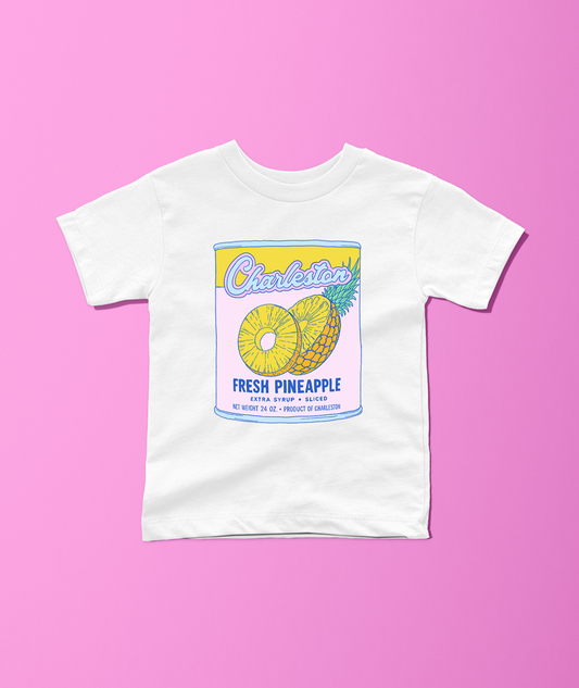 A white short-sleeve crewneck t-shirt featuring a colorful retro-style graphic of a pineapple can labeled 'Charleston Fresh Pineapple', displayed flat against a solid pink background.
