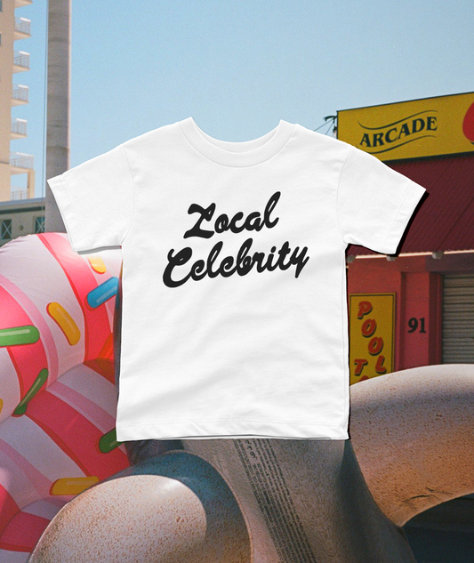 A white short-sleeve t-shirt with 'Local Celebrity' printed in black script across the chest, shown floating over a colorful outdoor street scene with a donut sculpture and storefronts.