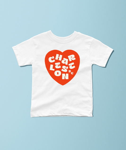A white short-sleeve crewneck t-shirt featuring a red heart graphic on the chest with the text 'CHARLES TON' in white stylized lettering, displayed flat against a light blue background.