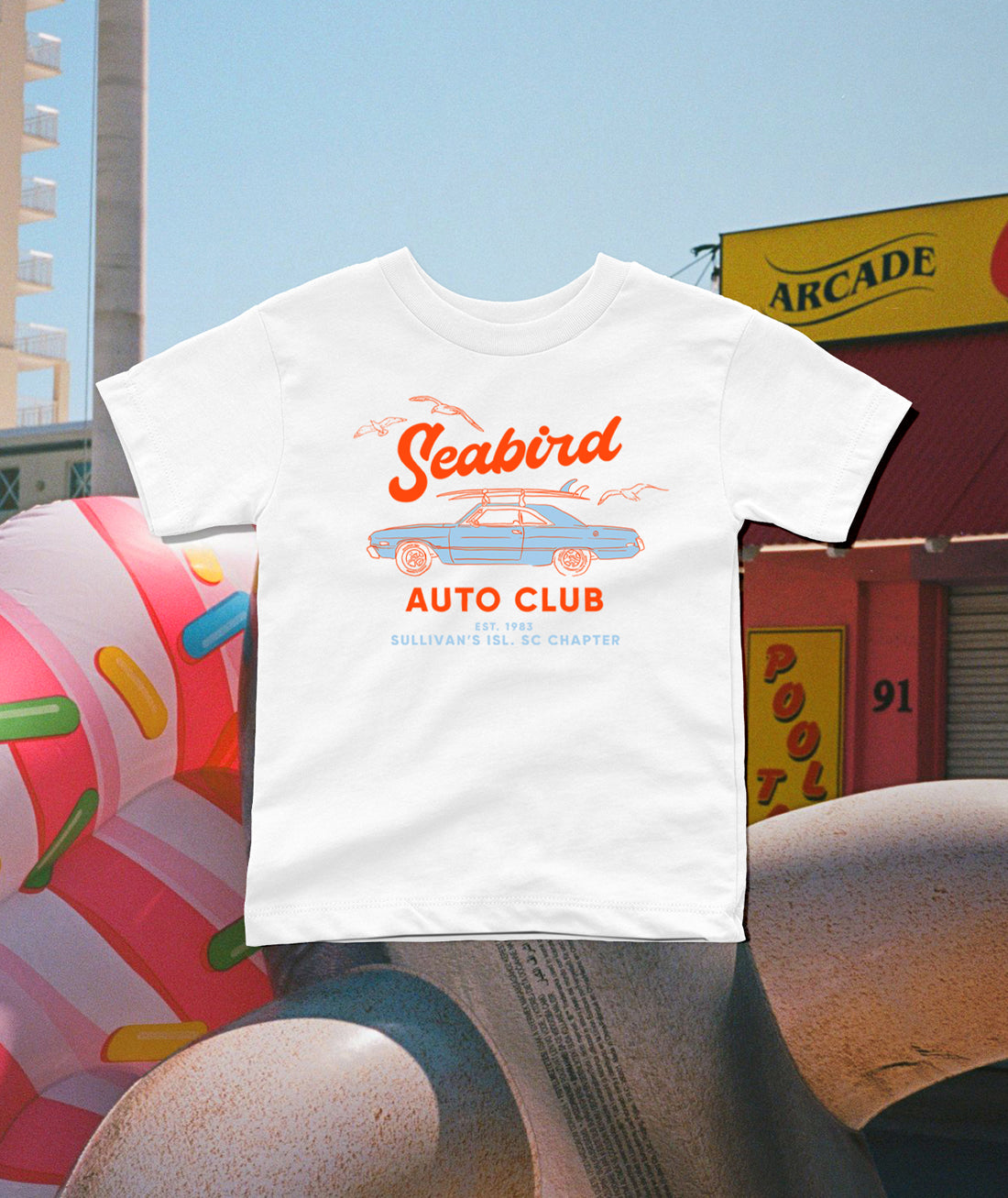 A white short-sleeve t-shirt with a retro orange and blue 'Seabird Auto Club' graphic, displayed in a stylized outdoor street scene with a donut sculpture and arcade building in the background.