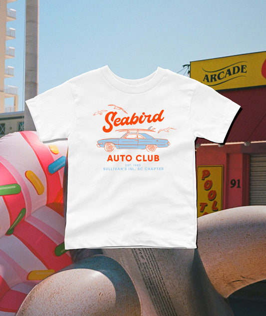 A white short-sleeve t-shirt with a retro orange and blue 'Seabird Auto Club' graphic, displayed in a stylized outdoor street scene with a donut sculpture and arcade building in the background.